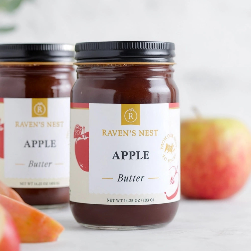Two glass jars of Raven's Nest Apple Butter next to a fresh apple and apple slices.
