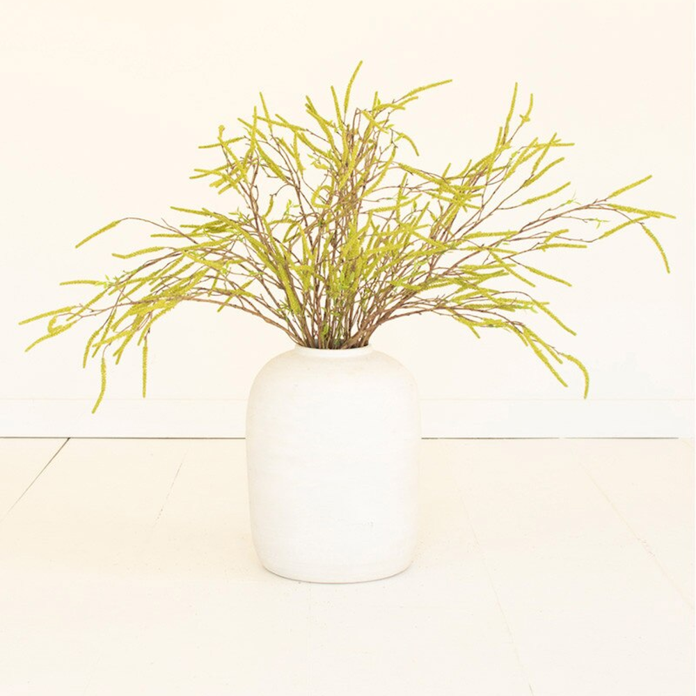 Artificial green willow flower sprays in a round white ceramic vase against a minimalist white background.