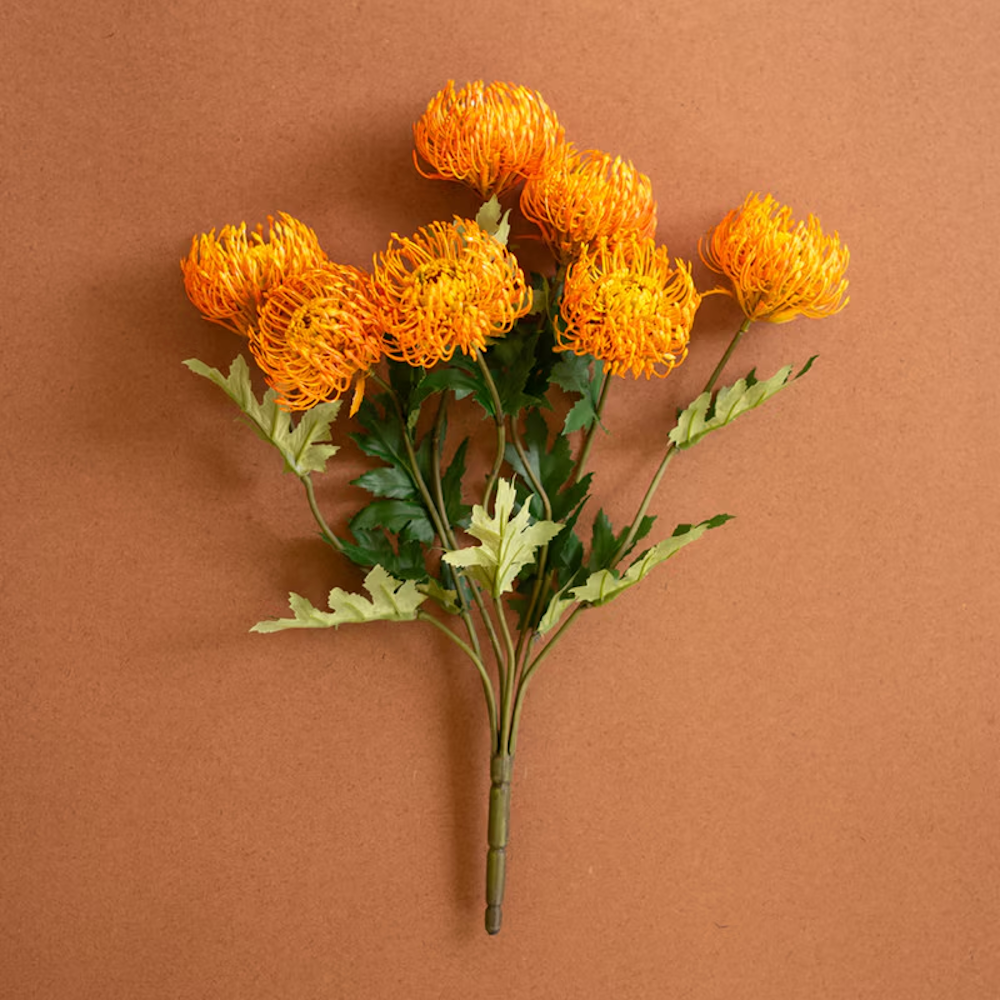 Artificial orange pincushion protea stem with green leaves on a warm brown background.