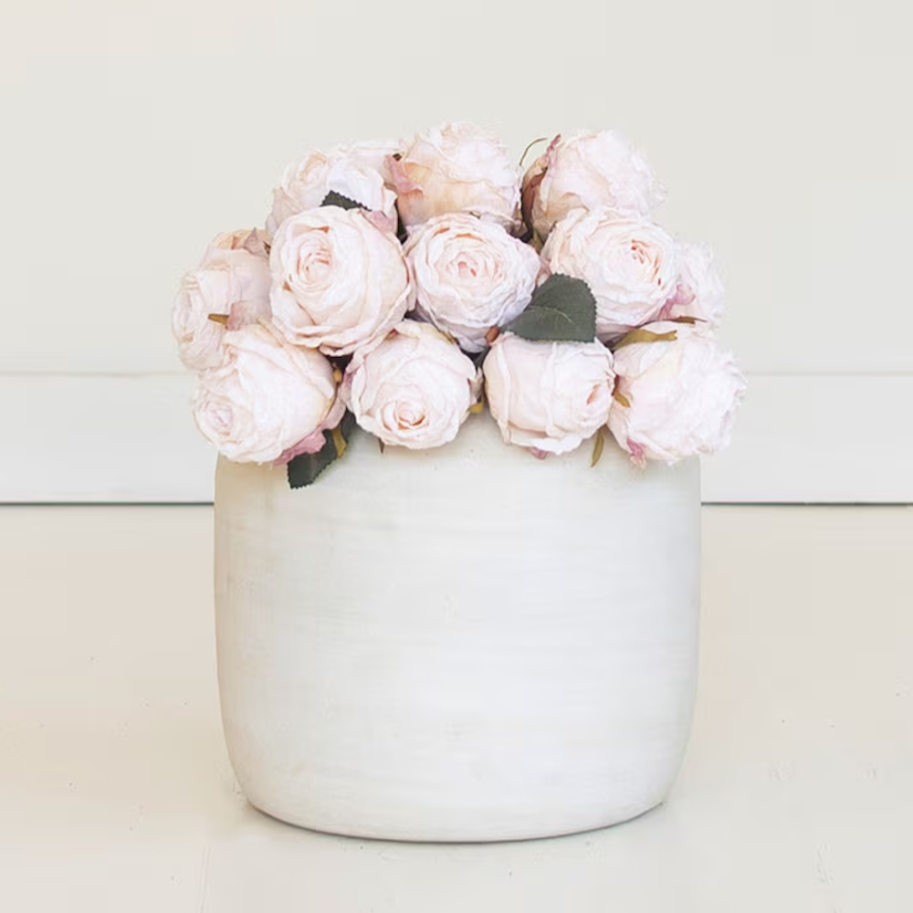 Soft blush pink artificial rose blooms arranged neatly in a white ceramic pot.