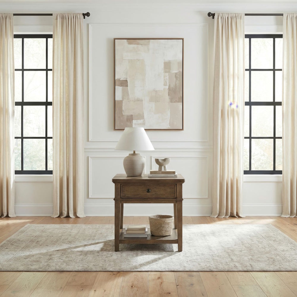Brown wooden end table with table lamp and decor in a bright room between two windows.