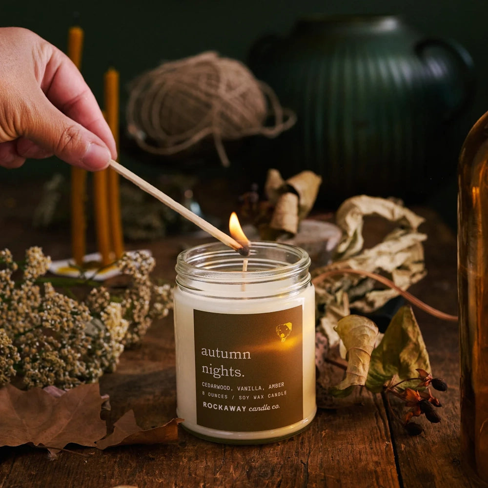 A hand lighting an Autumn Nights soy wax jar candle on a rustic wooden table with cozy fall decor.