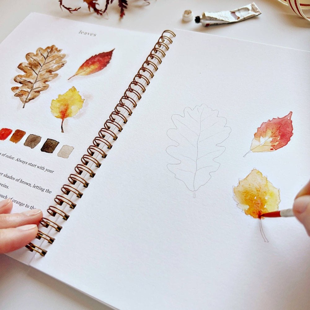 Open spiral sketchbook showing watercolor paintings and sketches of colorful autumn leaves.