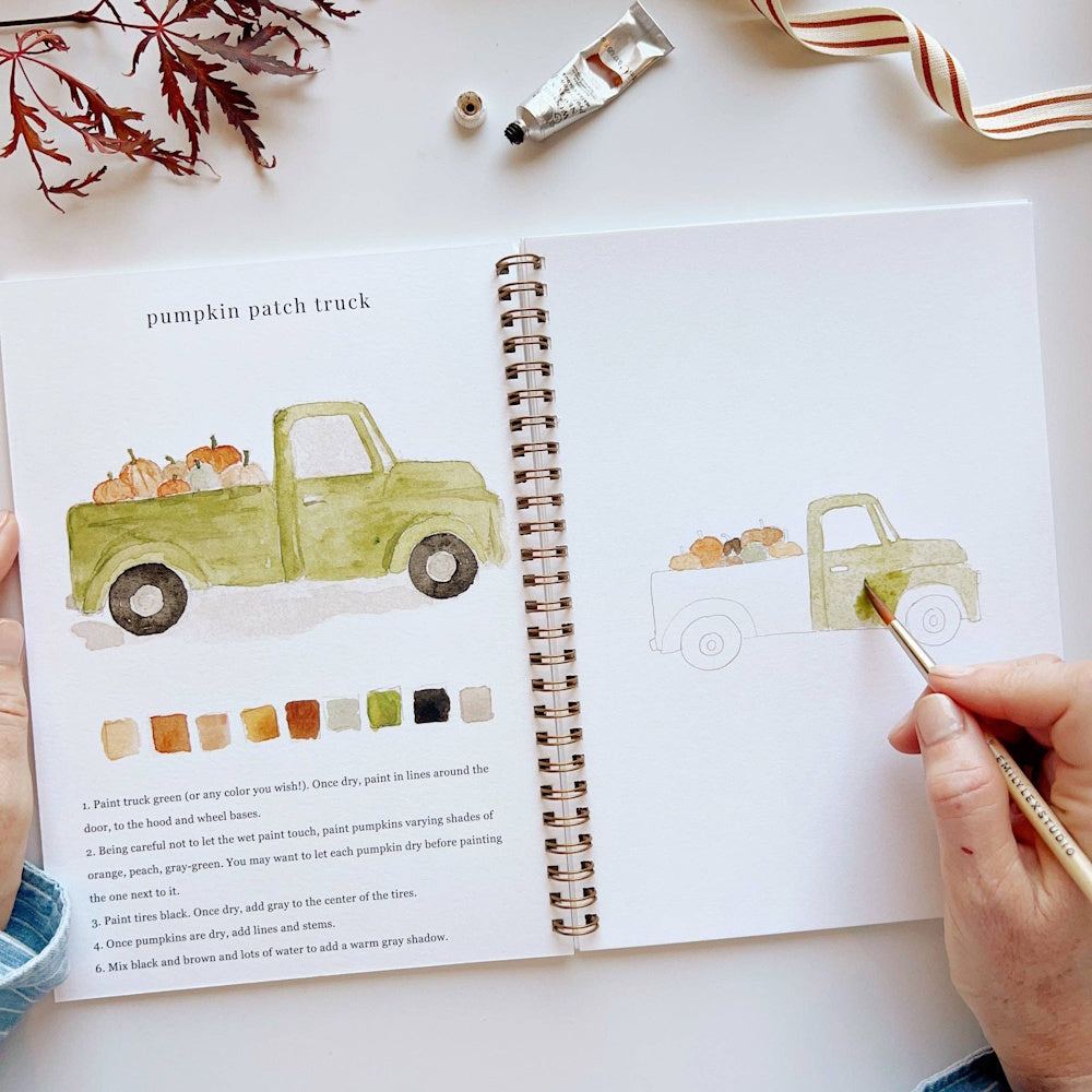Person painting a green pumpkin patch truck in a watercolor workbook with a paintbrush.