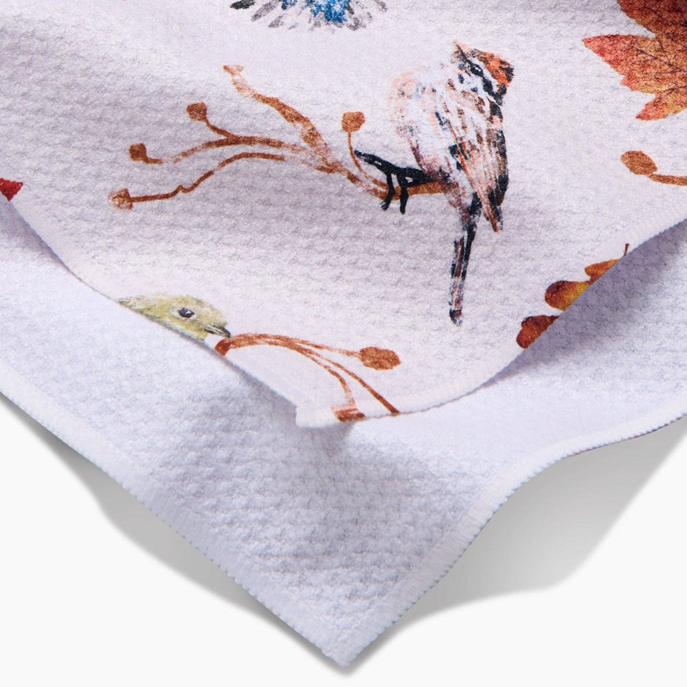 A folded white tea towel featuring a printed pattern of birds perched on branches with autumn leaves.