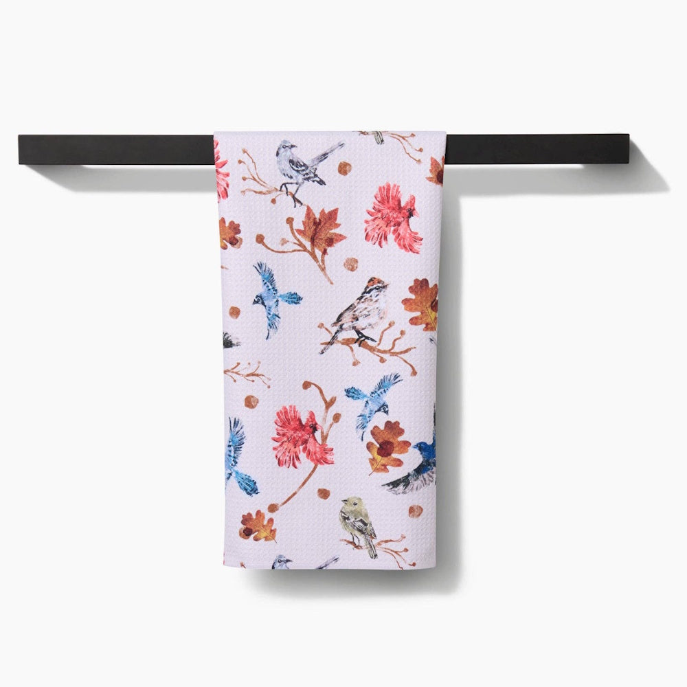 A textured tea towel featuring a pattern of autumn birds and fall leaves hanging from a black towel bar.