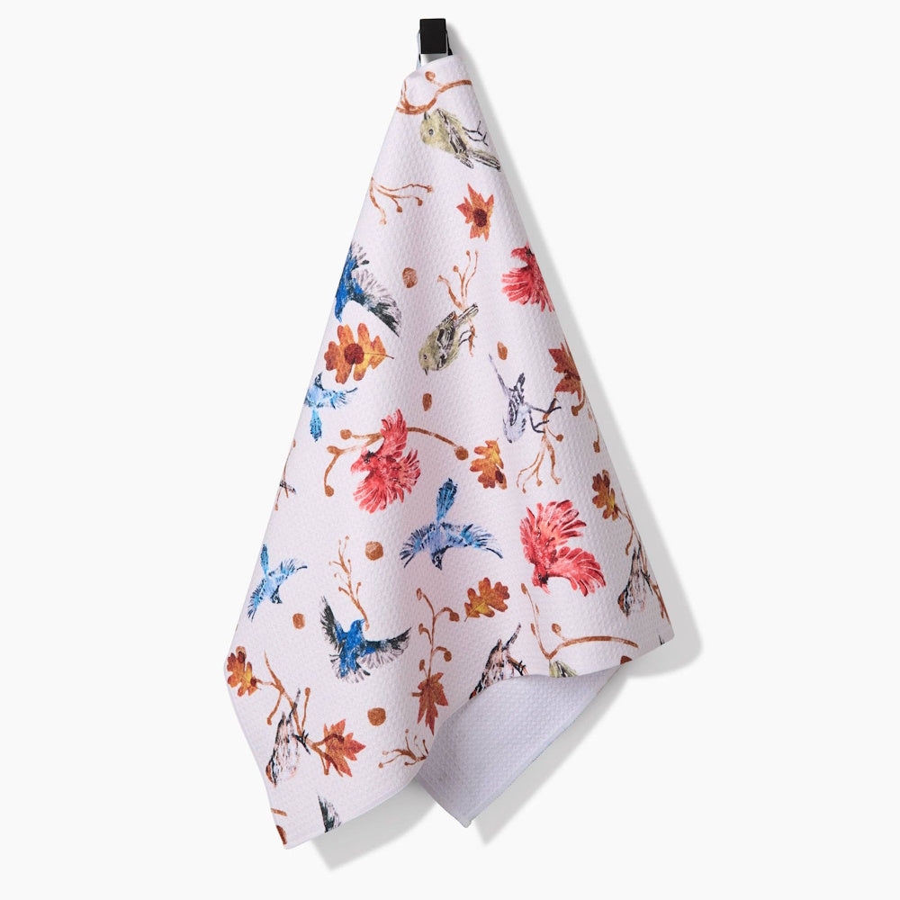 A white kitchen tea towel hanging up, featuring an autumnal pattern of blue, red, and brown birds and leaves.