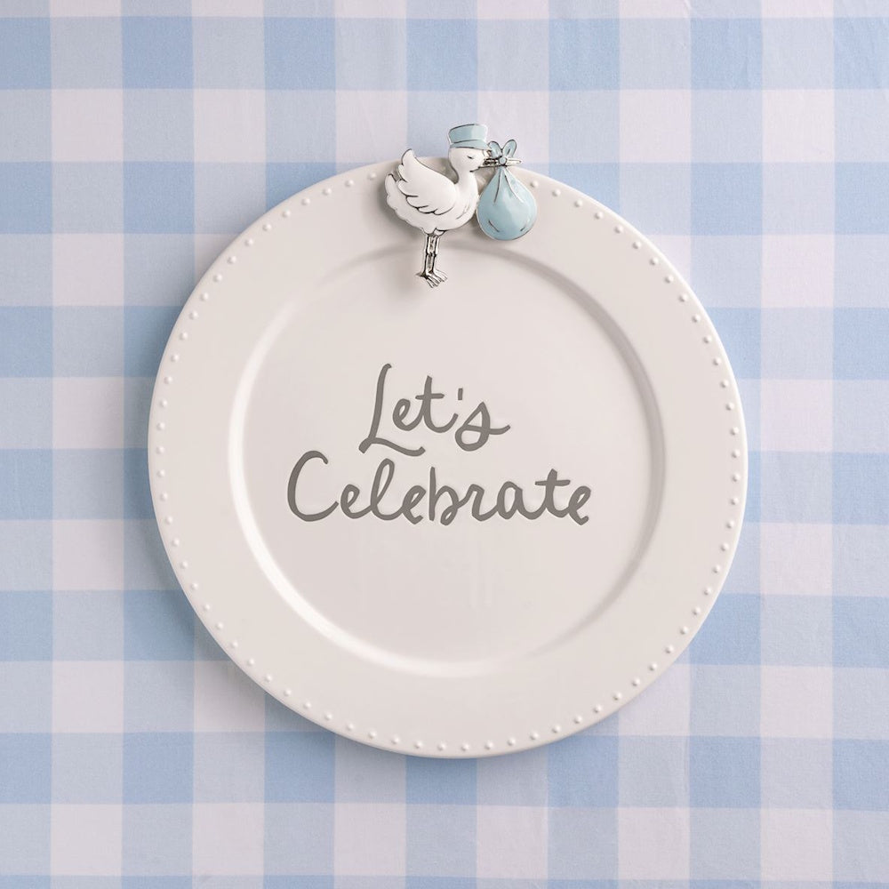 White "Let's Celebrate" plate with a baby boy stork charm on a blue checkered tablecloth.