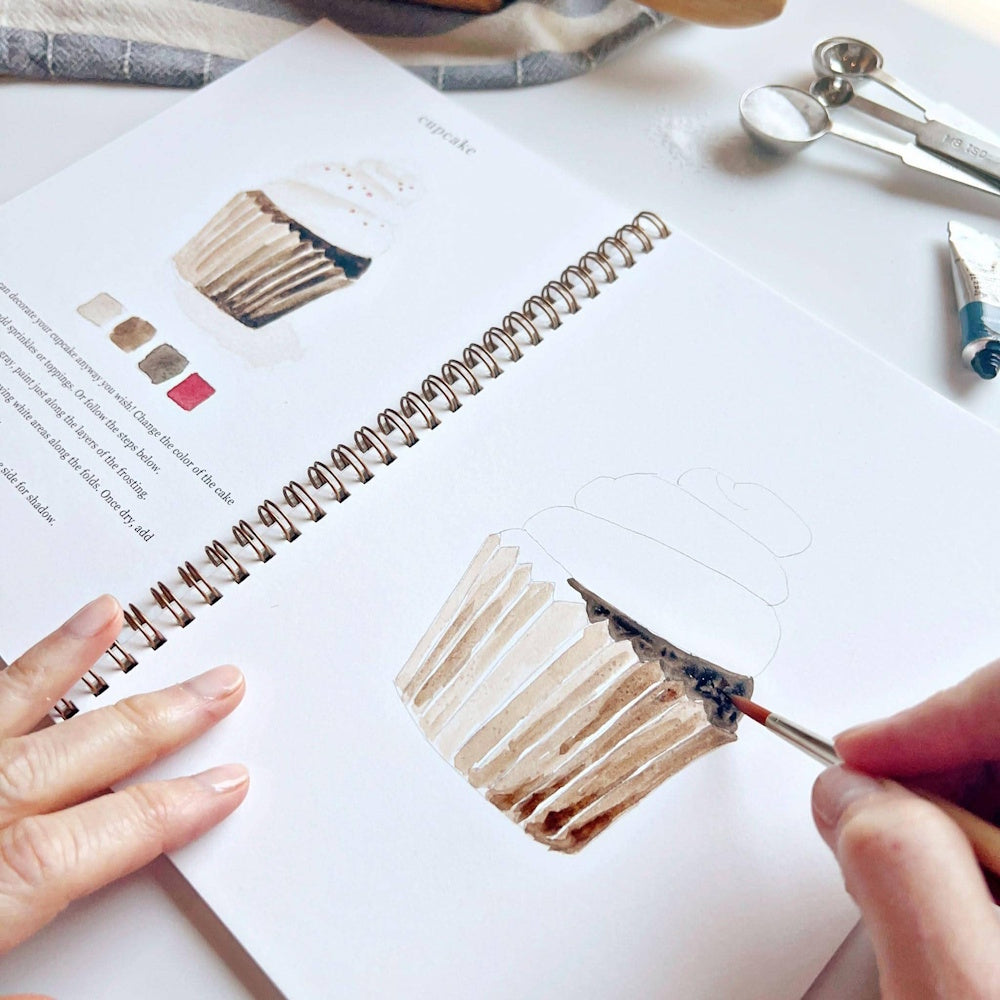A close-up of a person painting a cupcake in a watercolor workbook with brushes and paint supplies nearby.