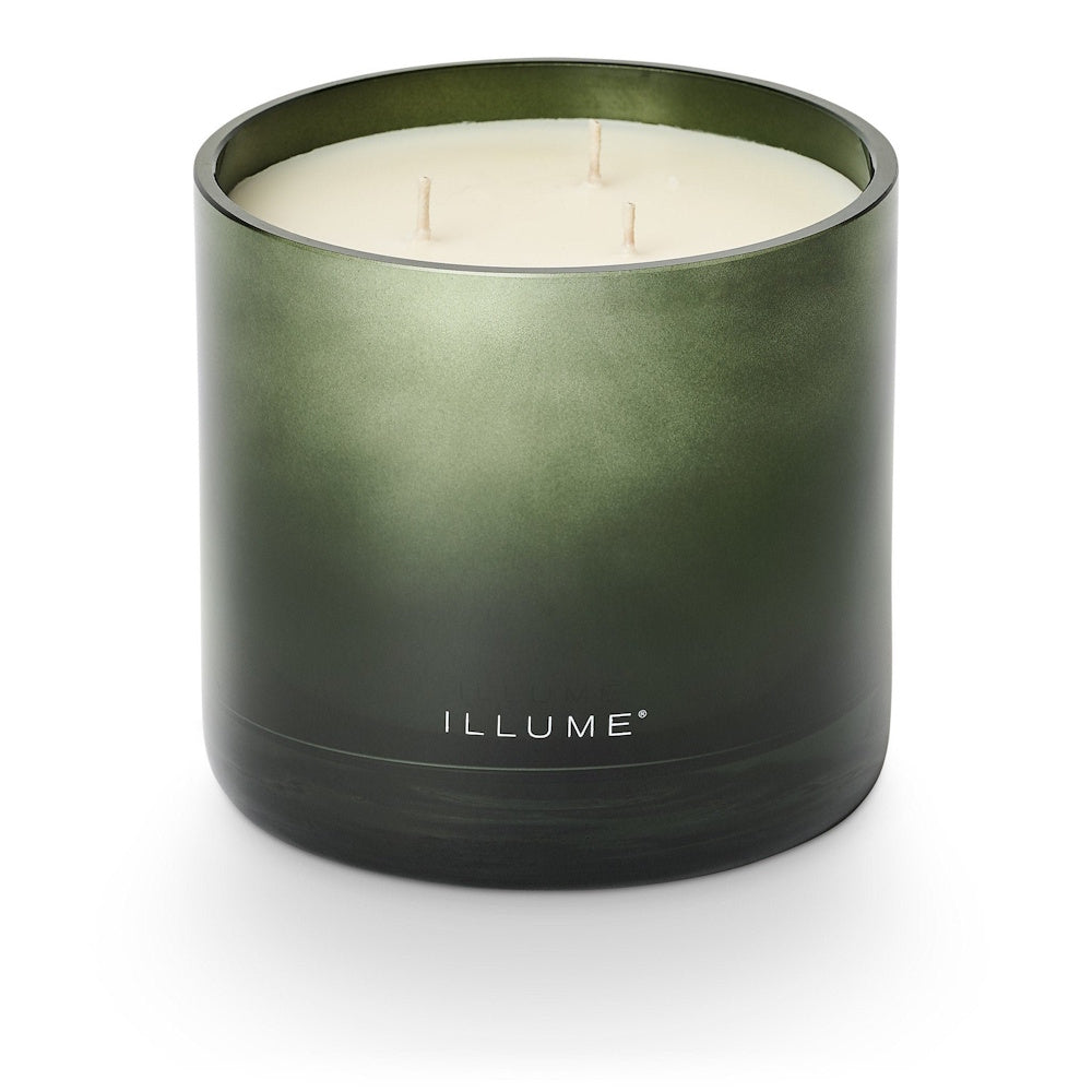 An unlit three-wick candle in a dark green glass jar with ILLUME branding.