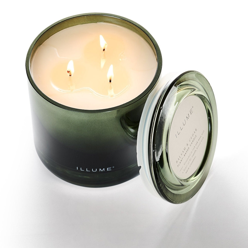 A three-wick lit candle in a dark green glass jar with its matching lid beside it.