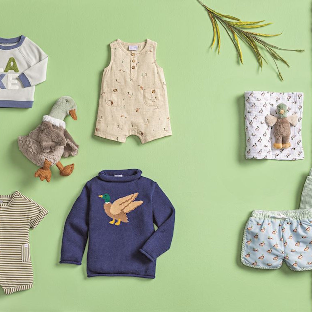Flat lay of mallard duck themed baby clothes, including a knit sweater, romper, and plush toys on a green background.