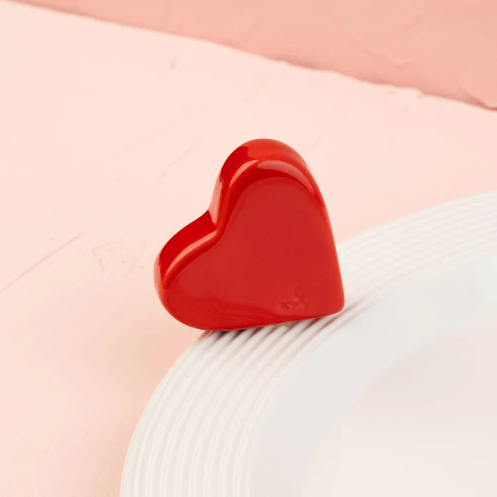 A glossy red ceramic heart figurine resting on the rim of a white textured plate against a pink background.
