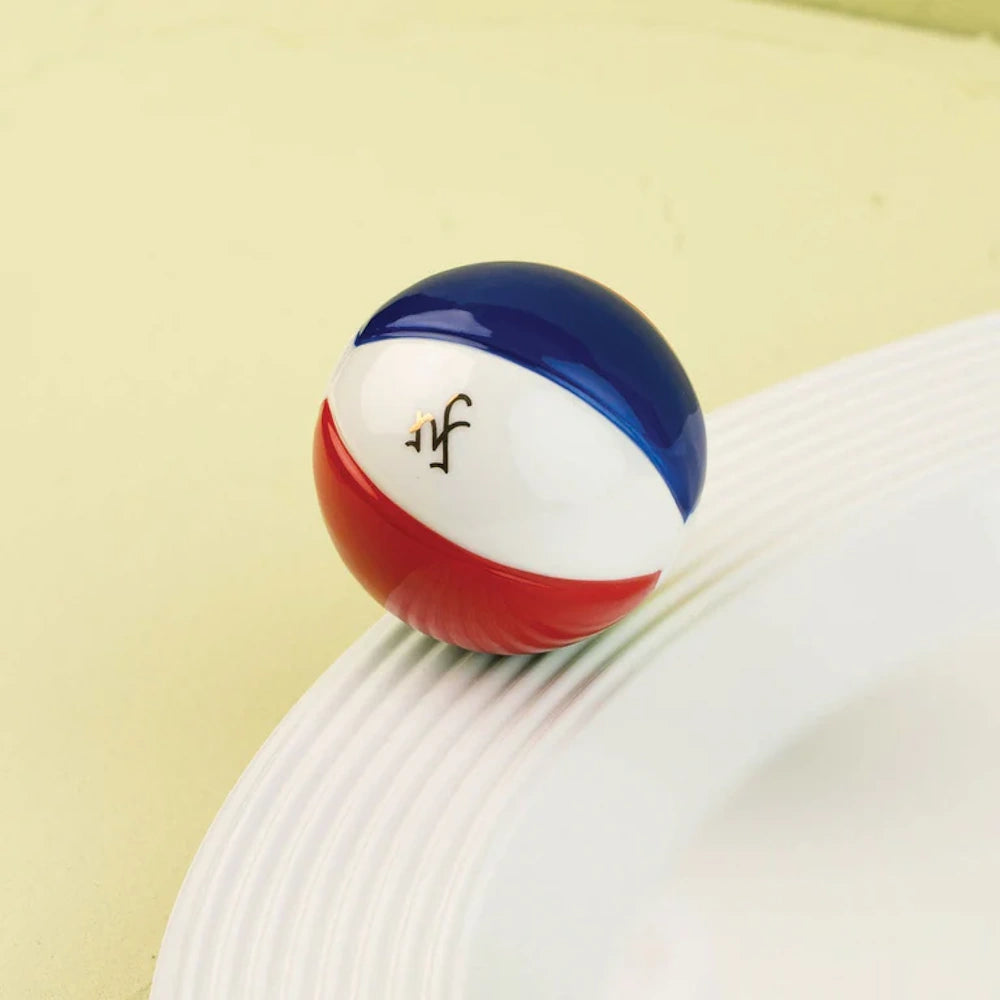 Small glossy ceramic beach ball charm in red, white, and blue attached to the rim of a white textured dish.