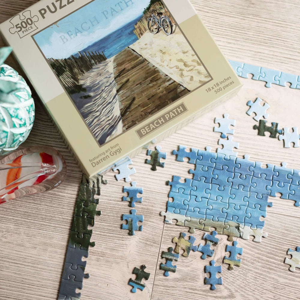 A partially assembled "Beach Path" jigsaw puzzle next to its box on a light wooden surface.