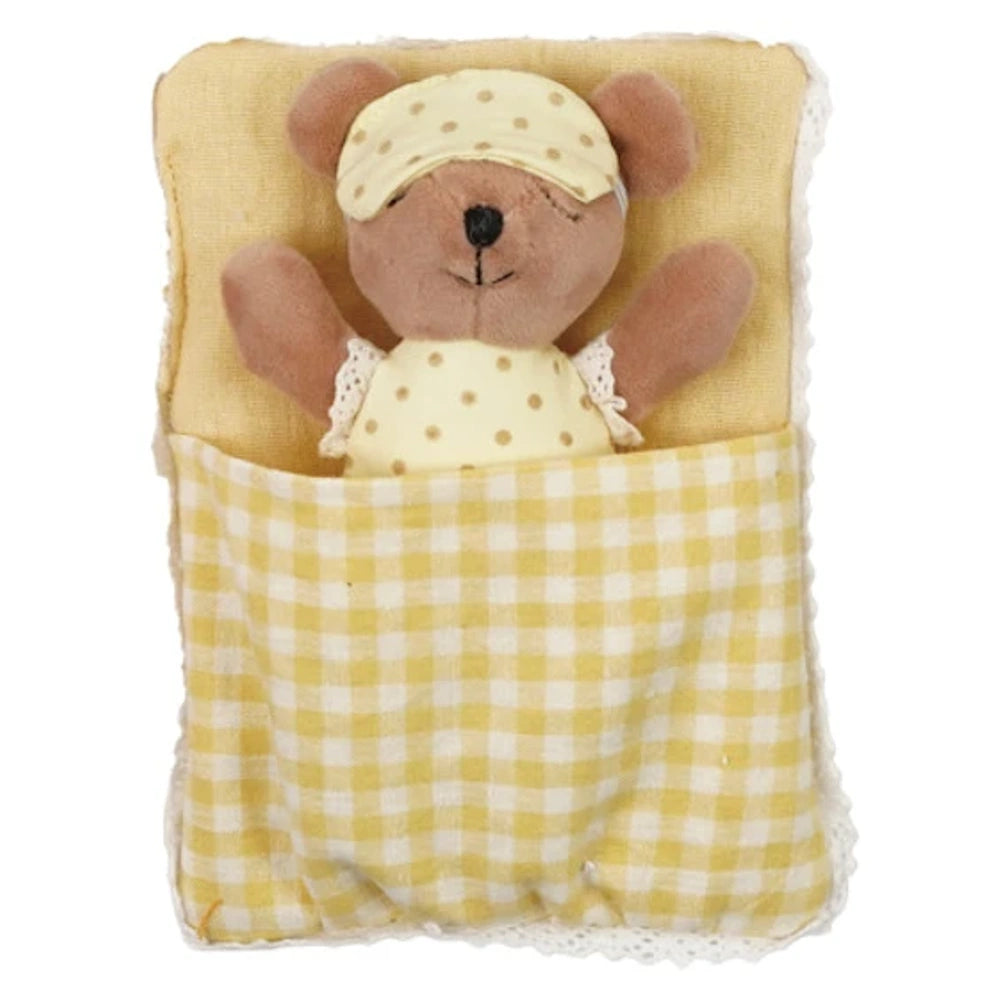 Small plush bear wearing a polka dot eye mask sleeping in a yellow plaid sleeping bag with lace trim.
