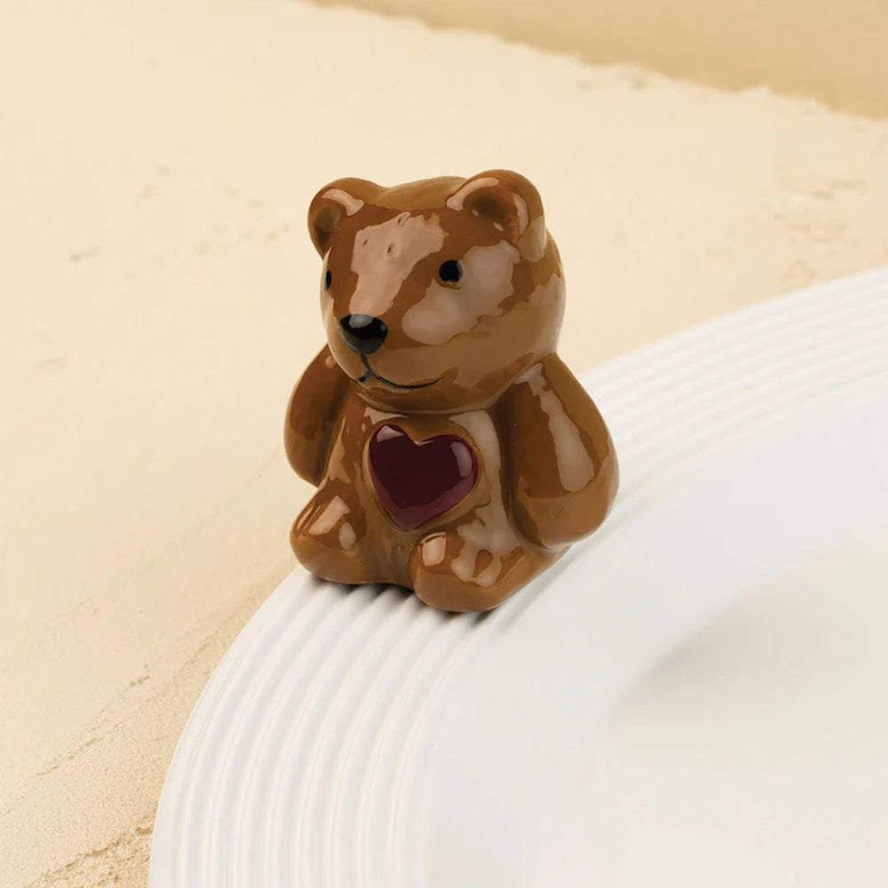 A small, glossy brown ceramic teddy bear with a red heart on its chest sitting on the rim of a white plate.