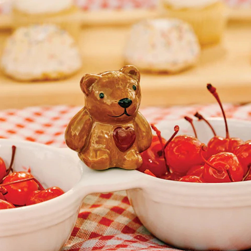 Brown ceramic teddy bear figurine with a red heart, sitting on a white serving dish filled with bright red cherries.