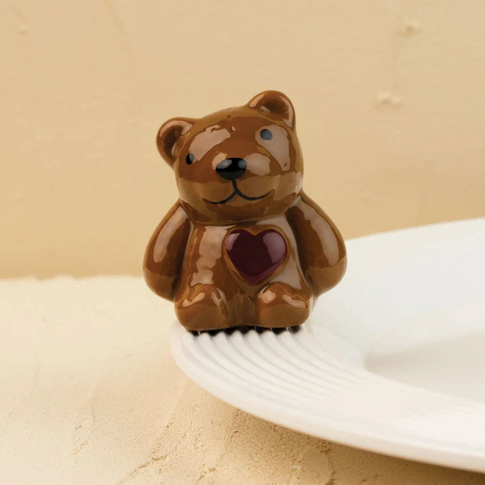 Glossy brown ceramic teddy bear figurine with a dark red heart on its chest sitting on the edge of a white plate.