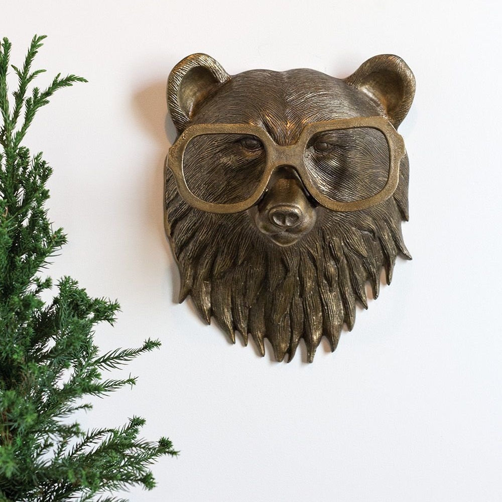 Bronze bear head wall sculpture wearing large glasses mounted on a white wall next to a pine plant.