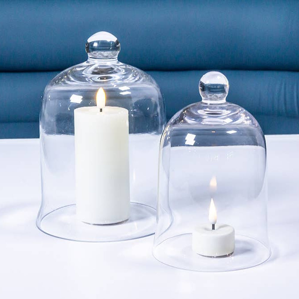 Two clear glass cloches with round knobs covering lit white candles on a white surface.