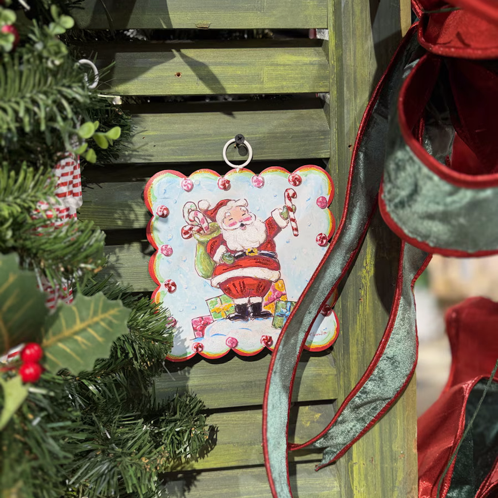 Lifestyle view of the Santa art plaque hanging against a green wooden shutter surrounded by holiday decor.