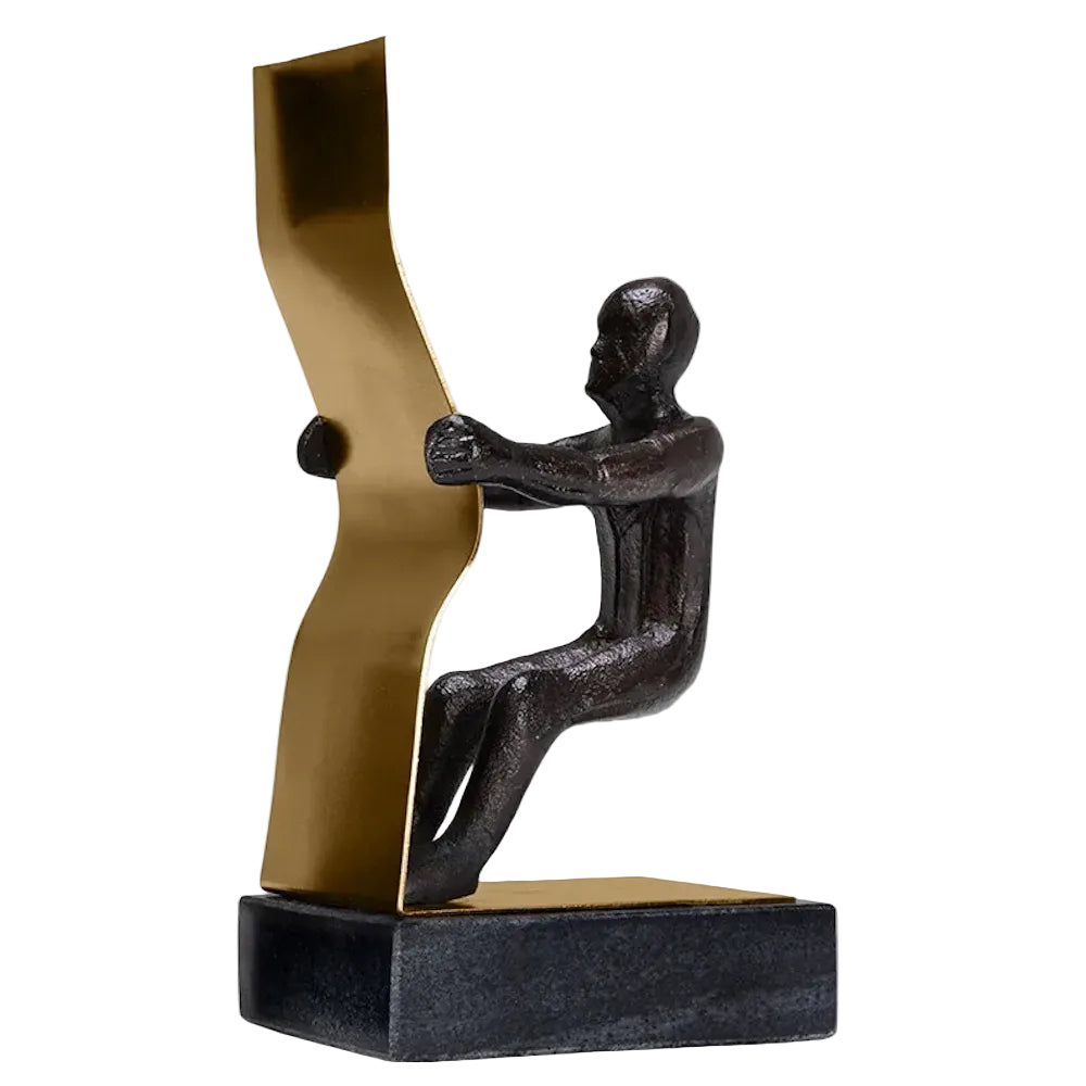 A dark metal figure sitting back and pulling on a curved gold metal ribbon, mounted on a black base.