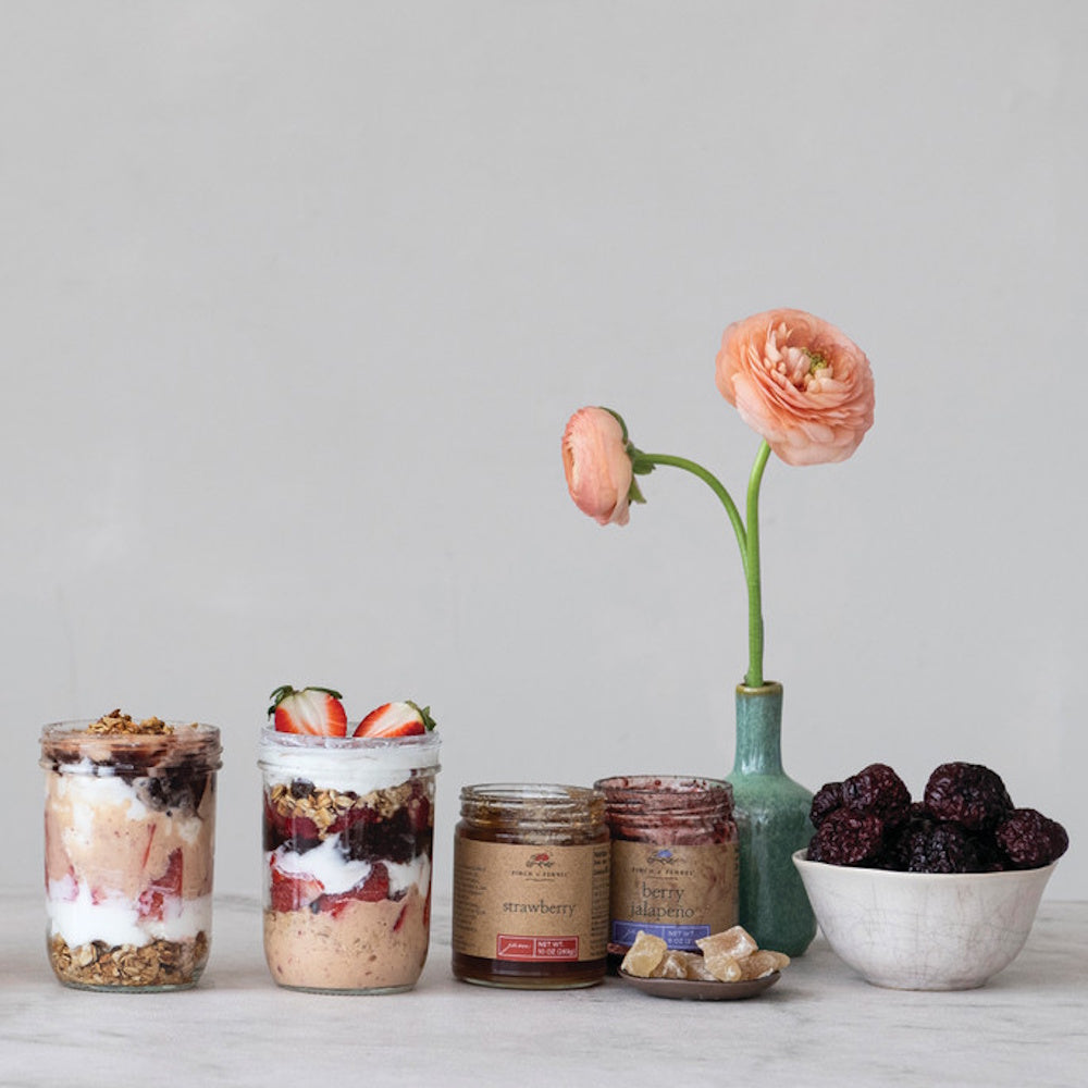 Jars of fruit parfaits, Finch + Fennel strawberry and berry jalapeño jams, fresh berries, and a pink flower.
