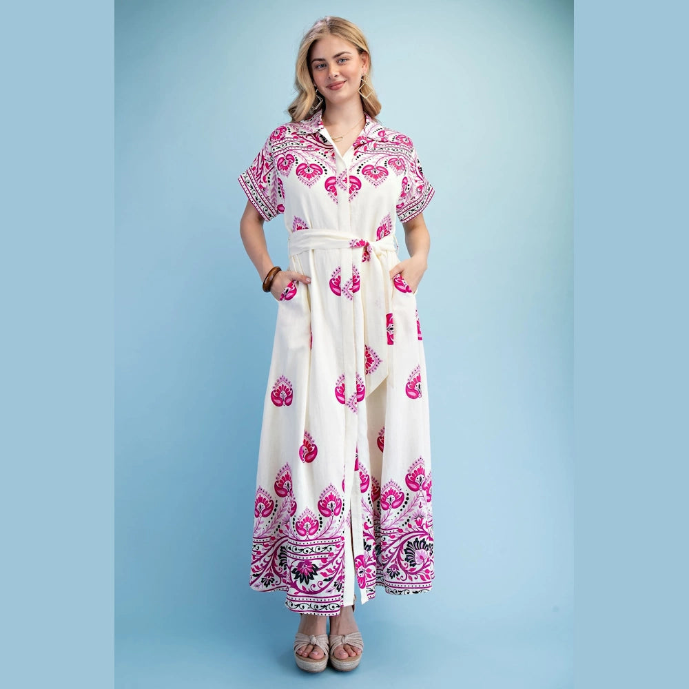 A woman in a white and pink patterned maxi shirt dress with short sleeves and a belted waist, standing against a blue background.