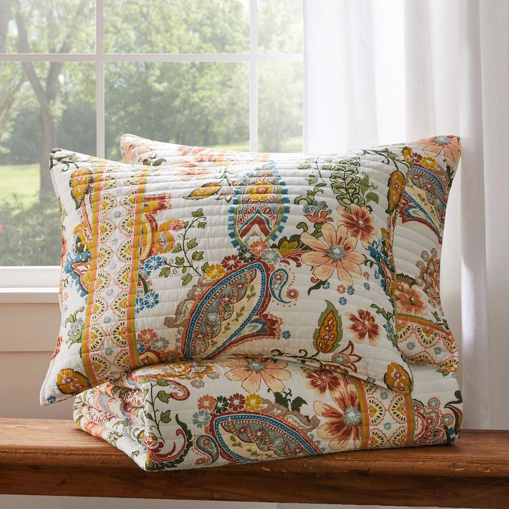 Two matching pillow shams with colorful paisley and floral patterns resting on top of a neatly folded quilt on a wooden bench by a window.