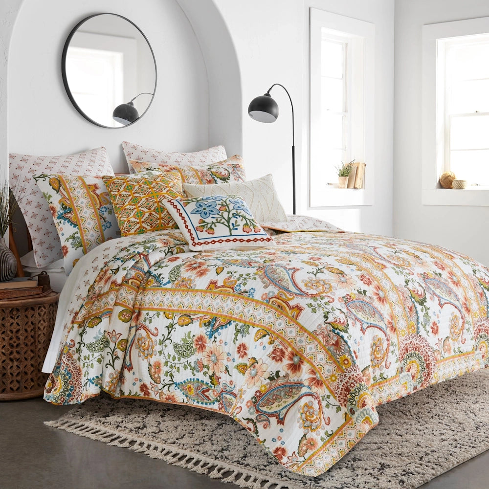 Side view of a bed styled with a colorful paisley quilt set, accent pillows, a black floor lamp, and a round mirror in a brightly lit bedroom.