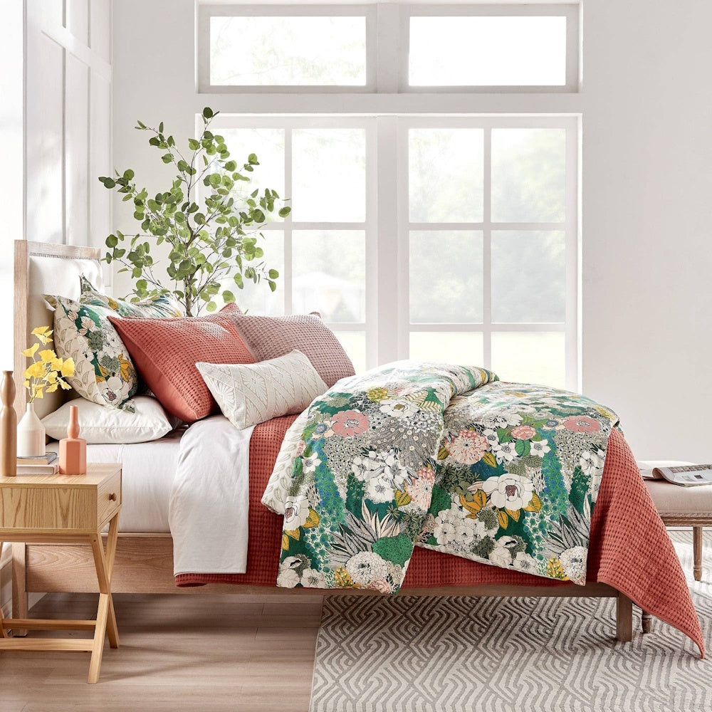 Floral comforter with terracotta throw blanket and pillows on a bed in a bright room with large windows.