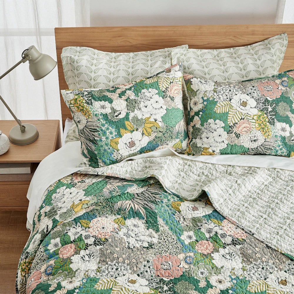 Floral quilt set with green, pink, and white botanical patterns on a wooden bed with a grey lamp.