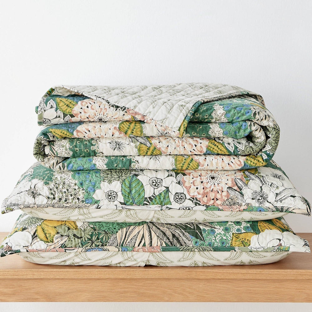 Stacked Bettina floral quilt and matching pillows with green, pink, and yellow botanical patterns on a wooden shelf.