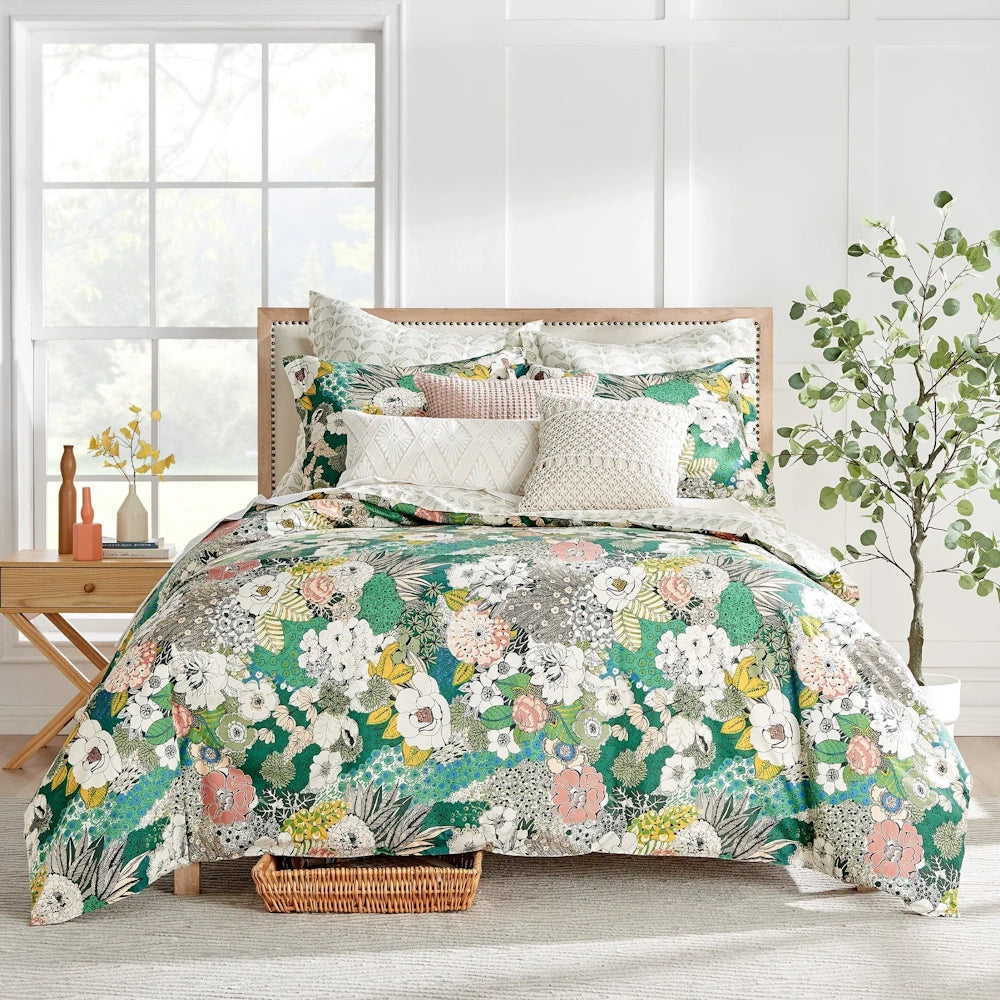 Floral bedding with green, pink, and white botanical patterns on a bed with decorative throw pillows in a bright room.