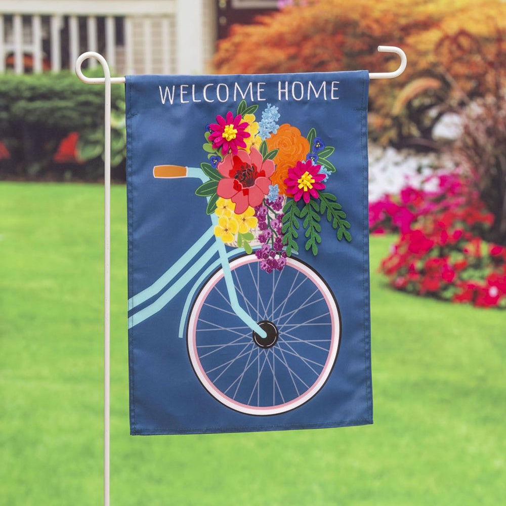 A blue garden flag with "WELCOME HOME" text and a 3D floral bicycle design, displayed on a lawn.