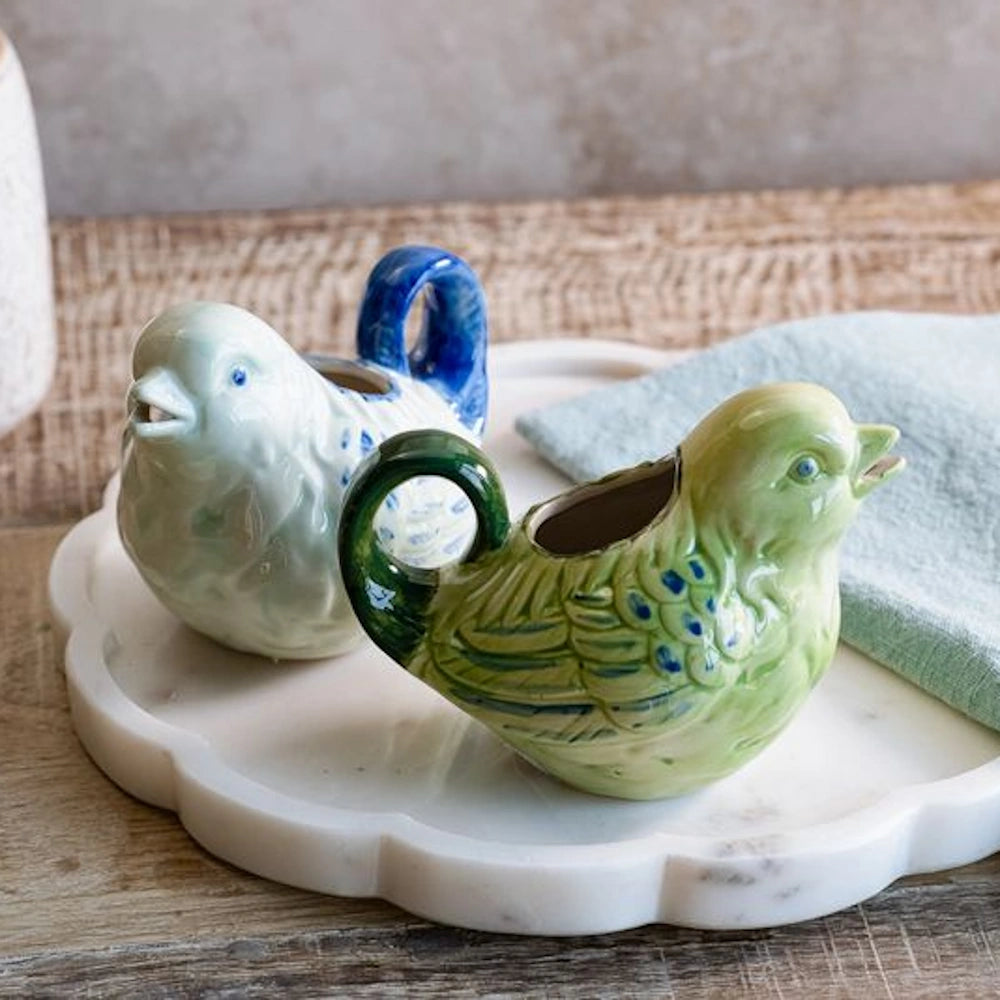 Two ceramic bird-shaped creamers, one green and one white with blue details, resting on a white marble scalloped tray.