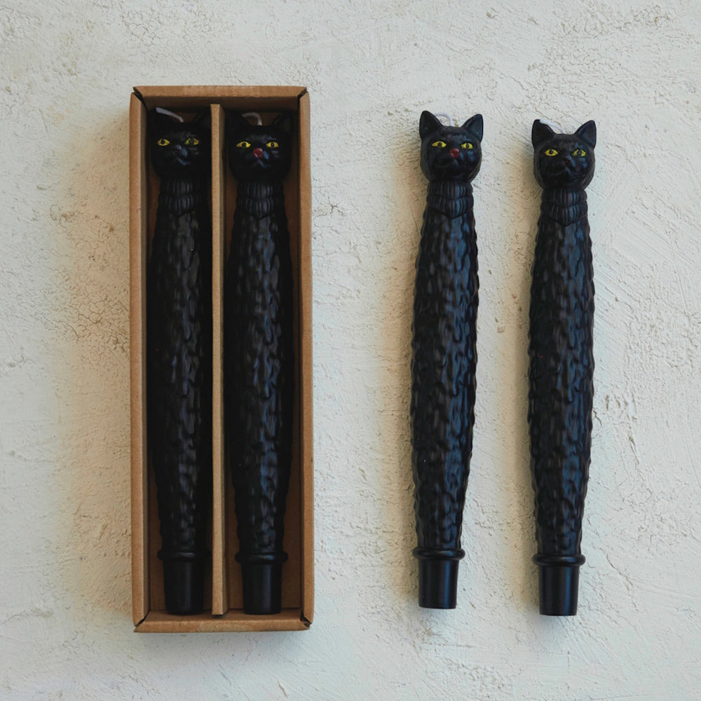 Two textured black cat taper candles in a cardboard box next to two loose ones on a white surface.