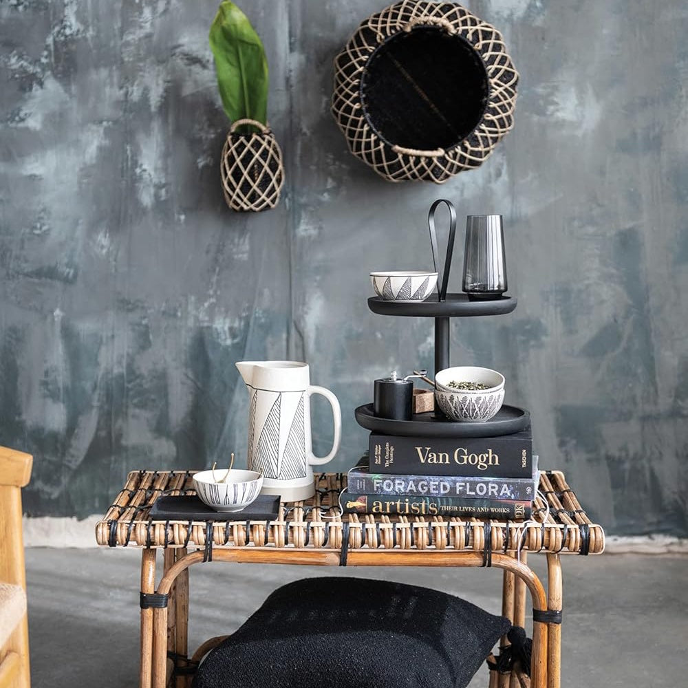 Stoneware pitcher, bowls, and tiered tray styled on a rattan table against a textured grey wall.