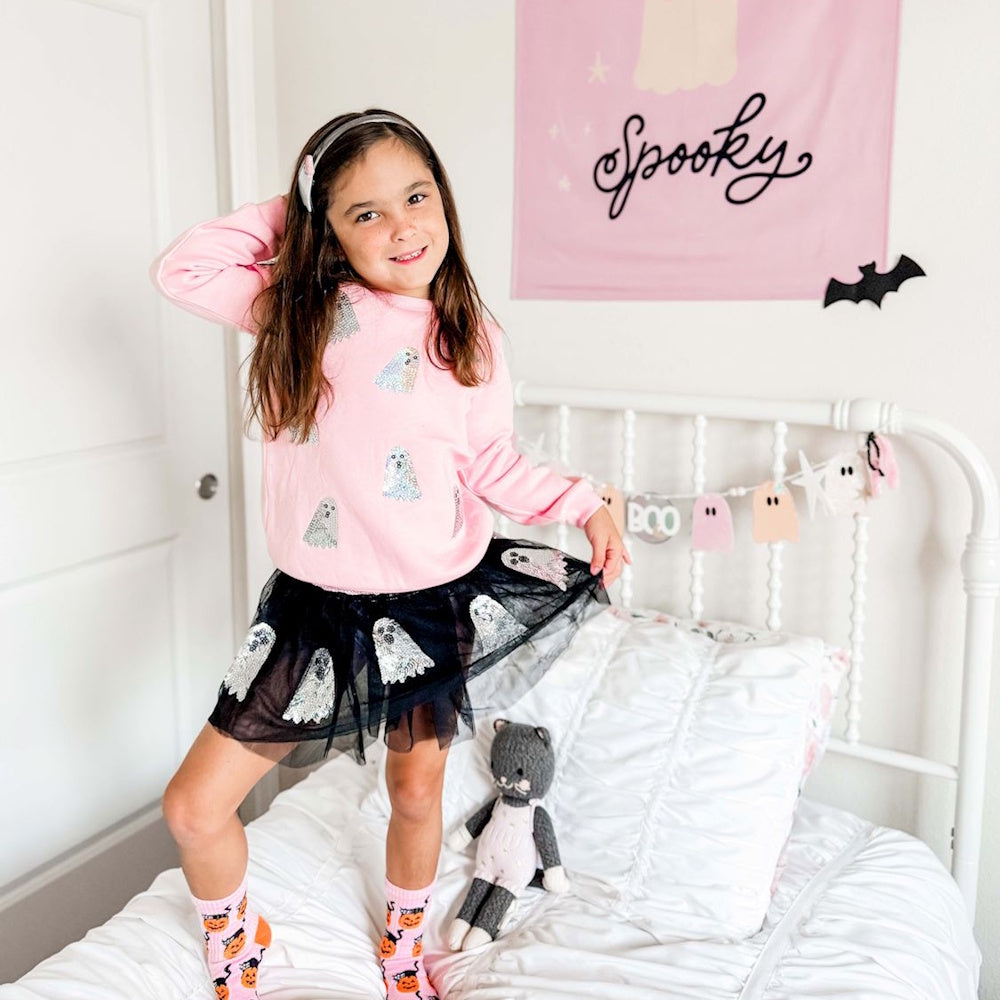 A young girl in a pink ghost sweatshirt and black sequin ghost tutu stands posing on a white bed.