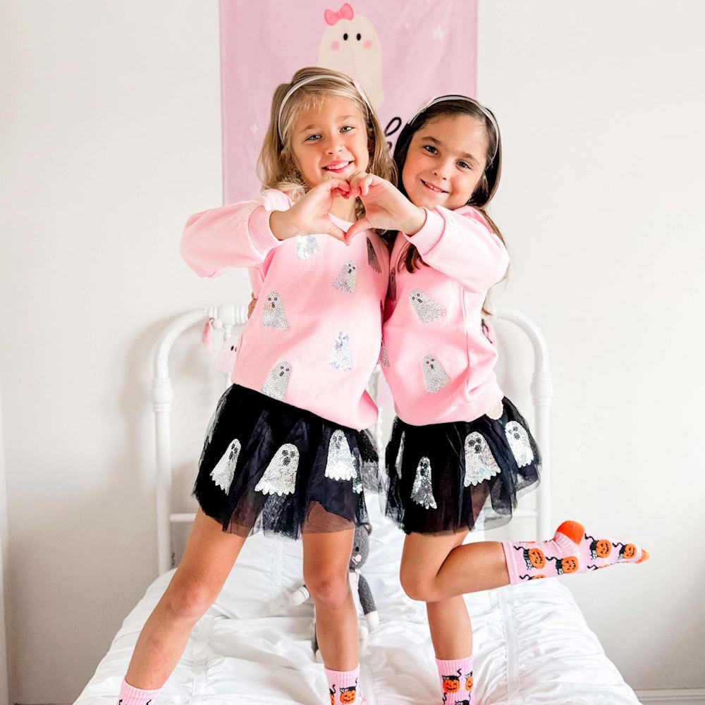Two girls wearing pink ghost sweatshirts and black sequin-ghost tutus make a heart shape with their hands.