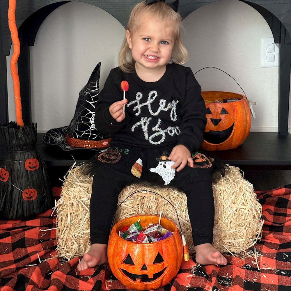 Smiling toddler in a 'Hey Boo' shirt sits on a hay bale surrounded by Halloween decorations and candy.