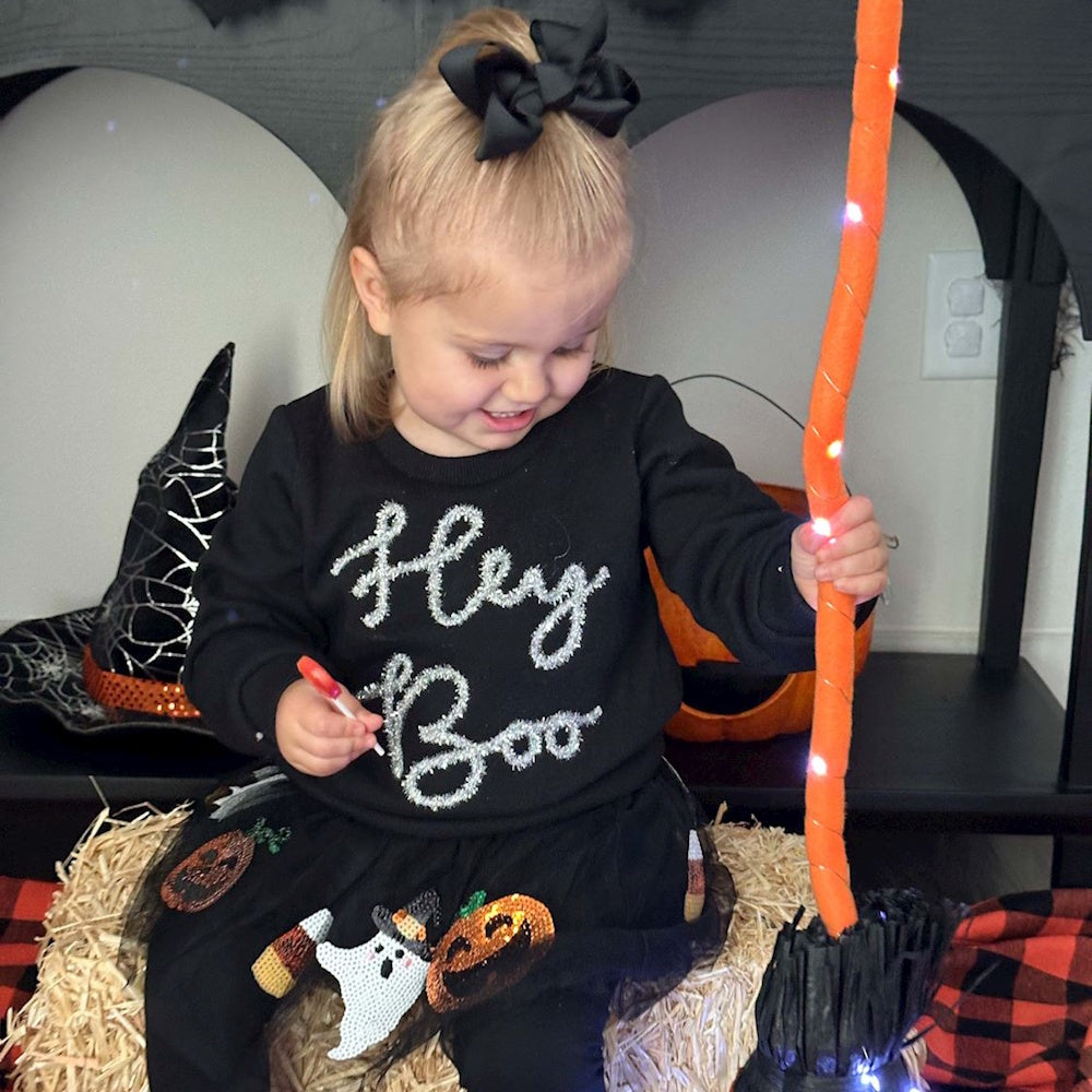A toddler in a "Hey Boo" sweatshirt sits on a hay bale holding a lollipop and a glowing orange broom.