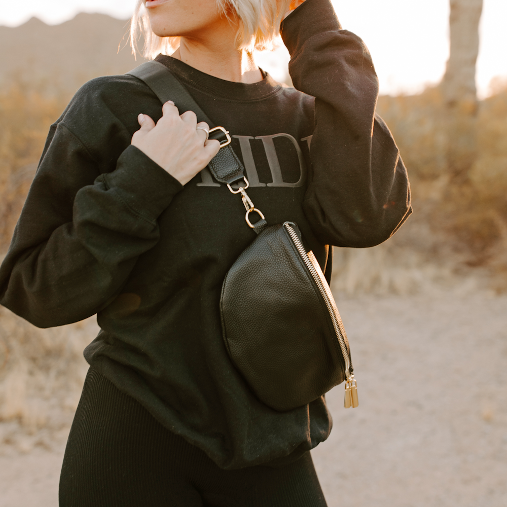 Woman wearing a black leather fanny pack as a crossbody bag over a black sweatshirt in a desert setting.