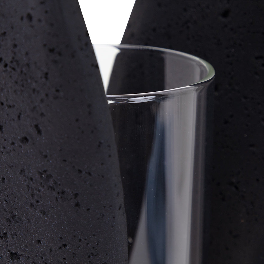 Close-up of a clear glass hurricane candle holder nestled between textured, porous black lava stone pillars.