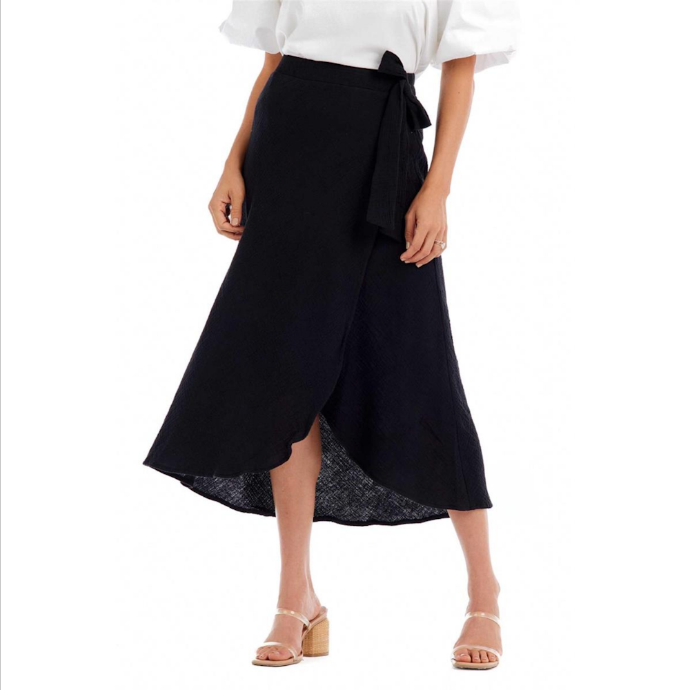 Woman wearing black midi-length wrap skirt with side tie waist, paired with white top and neutral heels.