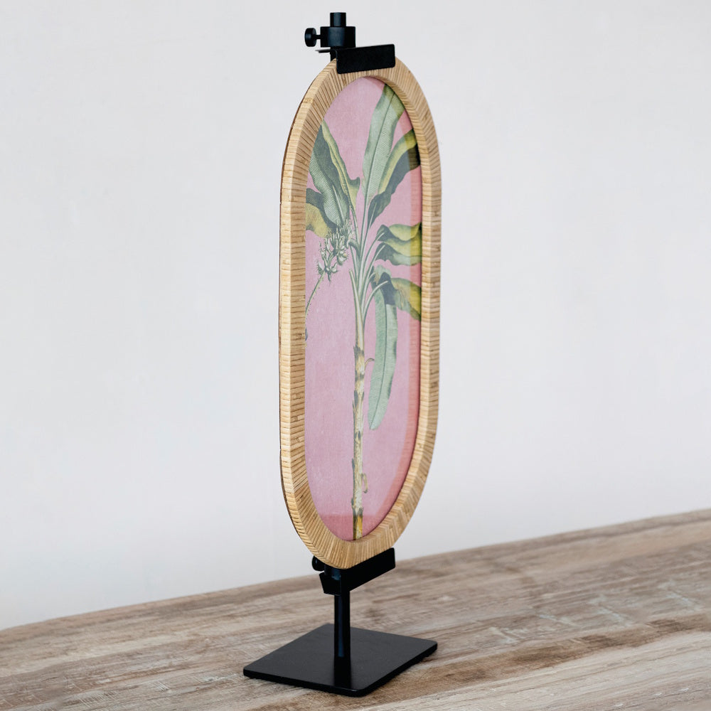 Oval tropical art print in a rattan frame displayed on a black metal adjustable tabletop easel.