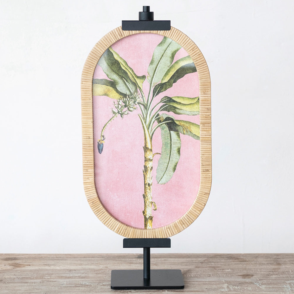 Tropical banana tree pink artwork in a rattan frame on a black metal adjustable tabletop easel.