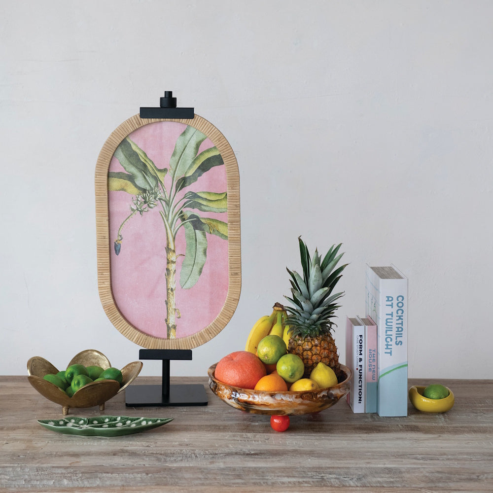 Tropical banana leaf art on stand with a fresh fruit bowl and books on a wooden table.
