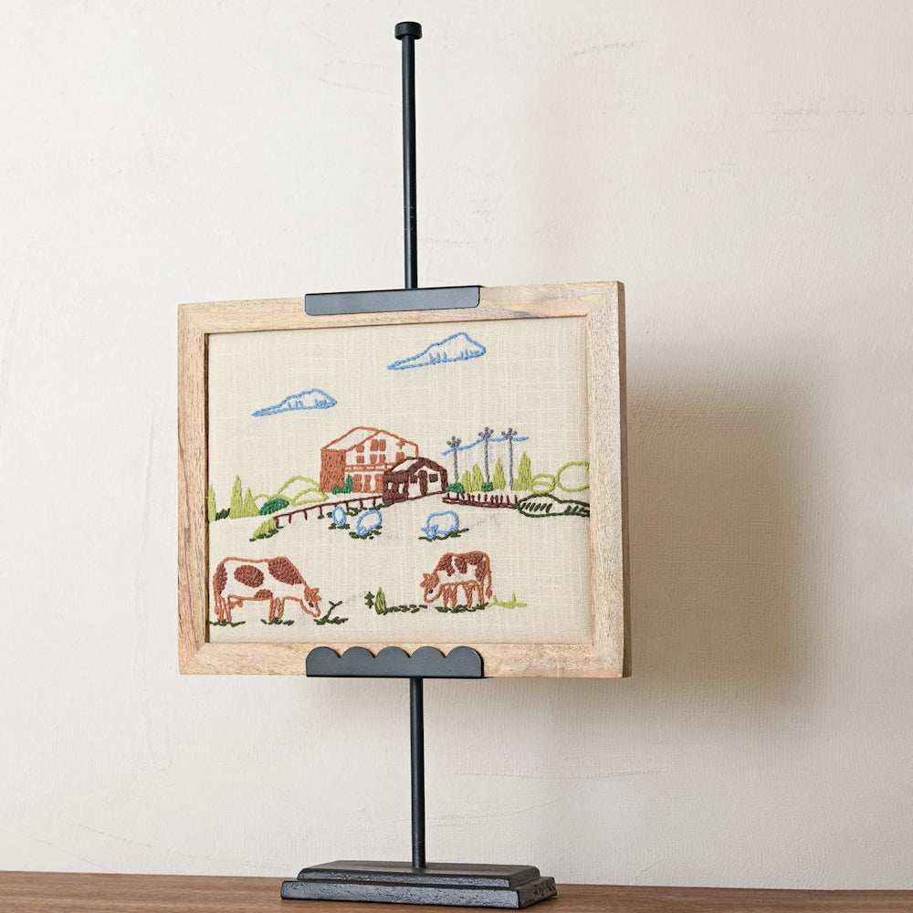 Black metal adjustable easel holding a framed rural farm embroidery with cows, a barn, and a scalloped base.